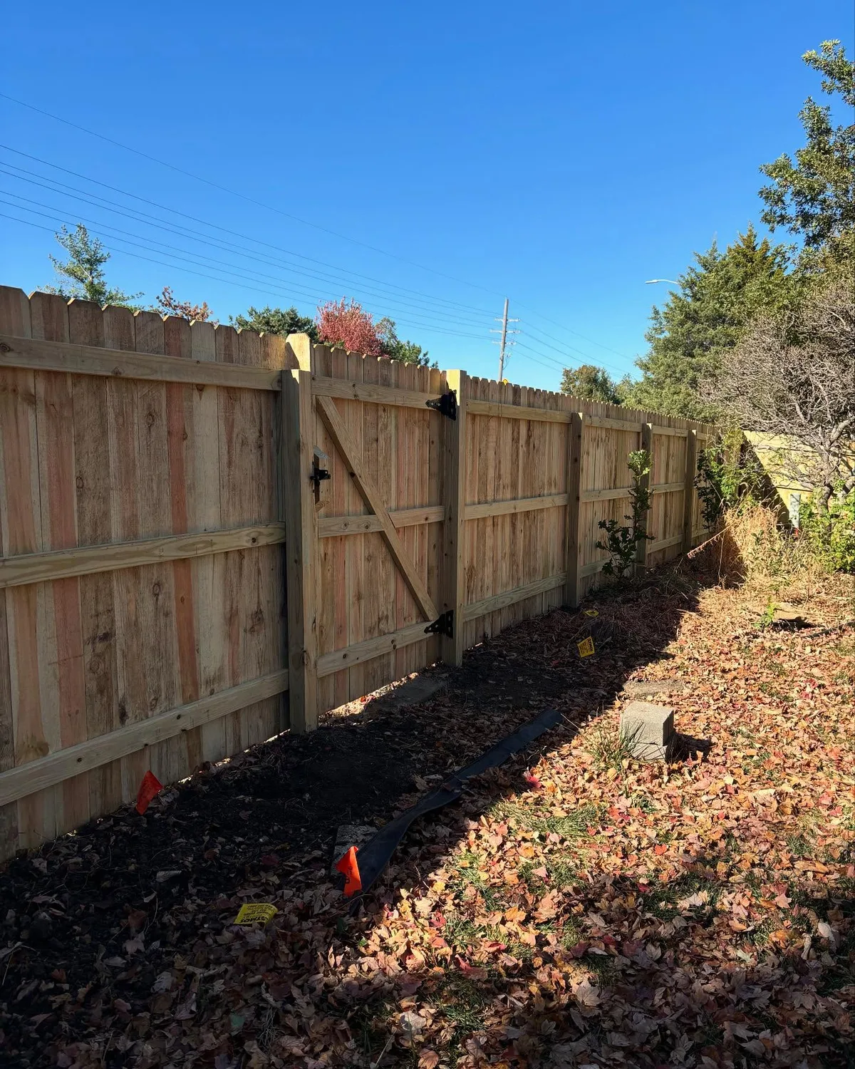 Remaining standing fence panels beside downed section in Lee's Summit, MO — fall tree line in background